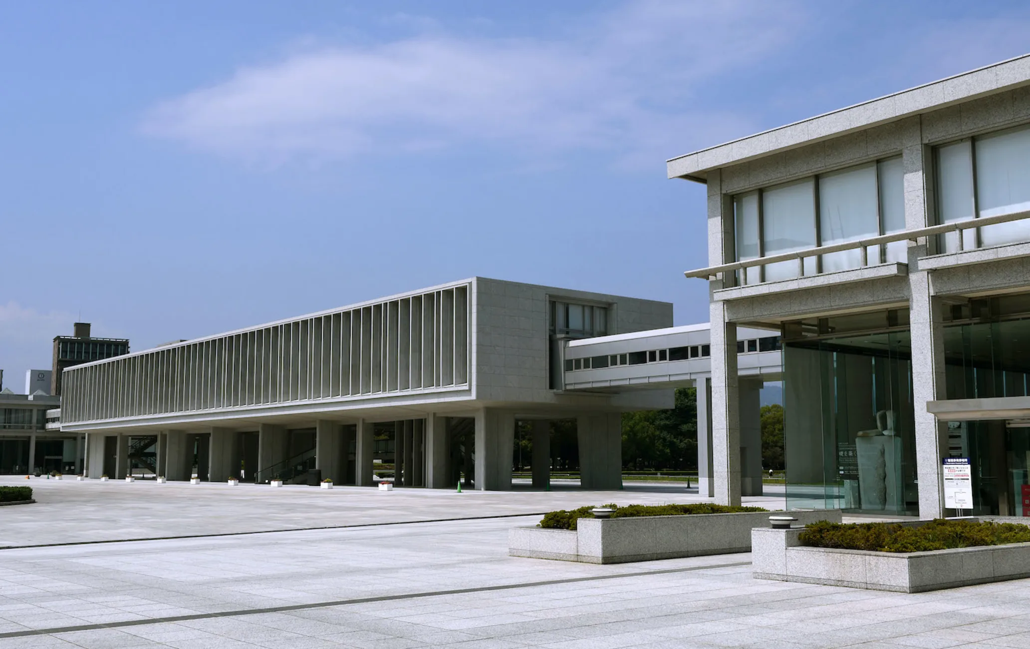 The grey concrete and glass buildings of the Hiroshima Peace Memorial Museum. A dark green tree can be seen in the background, and it is a sunny day with a clear sky.