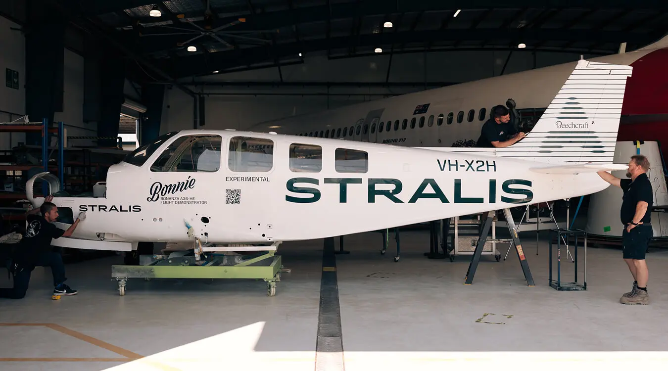A white Stralis Beech A36 Bonanza VXH-X2H jet sat in an aircraft hangar. It is being cleaned and inspected by staff dressed in black t-shirts and shorts.