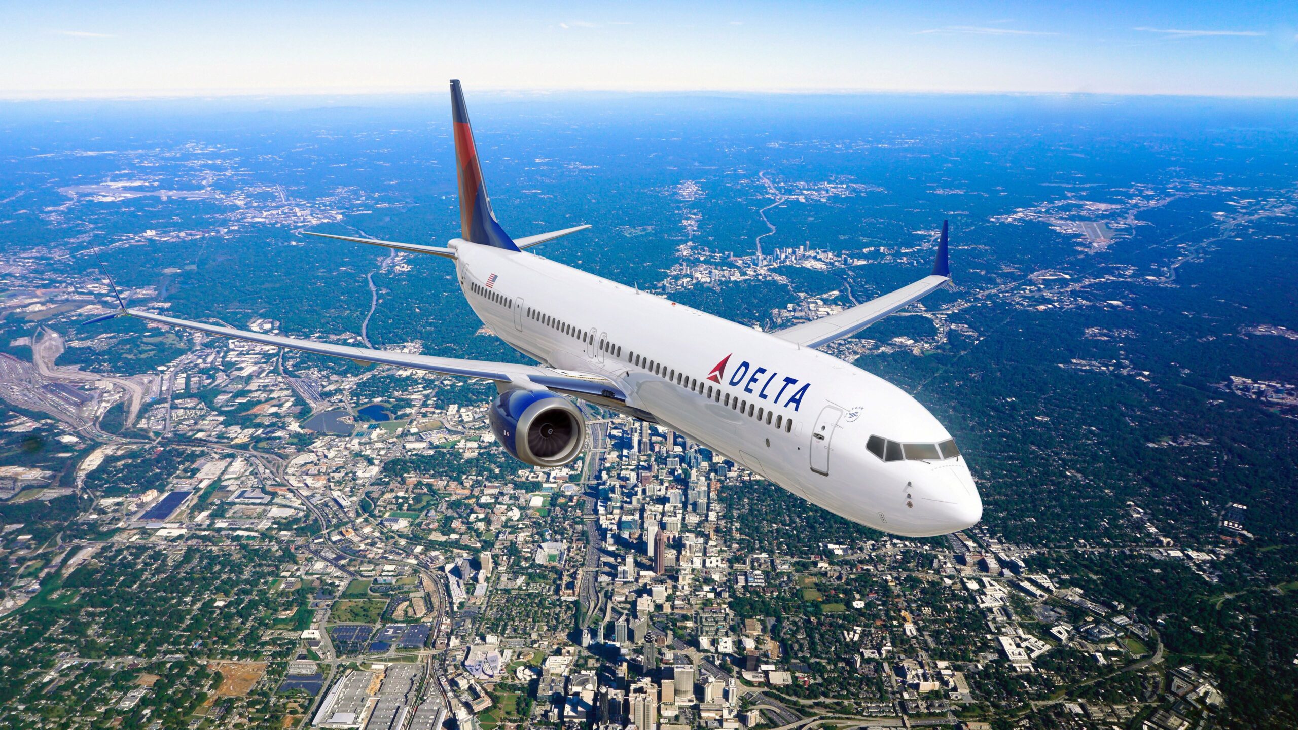 A red and white Boeing 737 Max 10 jet painted in the colours of Delta Air Lines. The plane is flying over a town, in front of a clear blue sky.