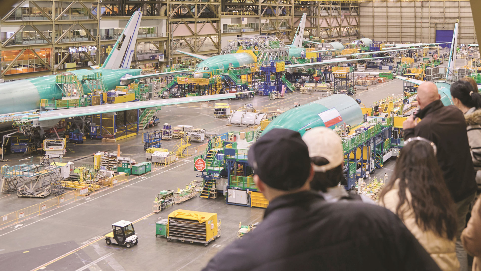 A group of people lean over a railing to observe work at a Boeing factory. Several white and green jets are pictured being constructed.