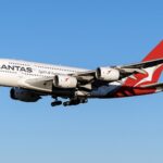 A red and white Airbus A380 jet in the livery of Qantas Airways. The jet is flying in front of a clear blue sky.