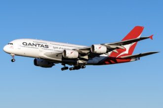A red and white Airbus A380 jet in the livery of Qantas Airways. The jet is flying in front of a clear blue sky.