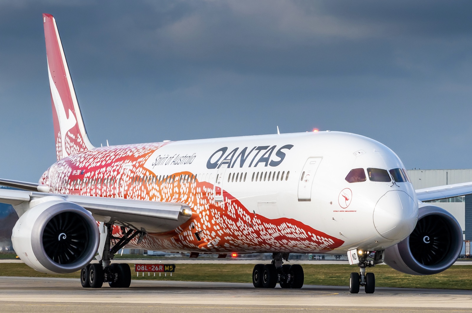 A red, orange, and white Boeing 788-9 jet sat on a runway. The aircraft has the livery of Qantas Airways painted on it, and is shown on a runway, in front of a grassy verge.