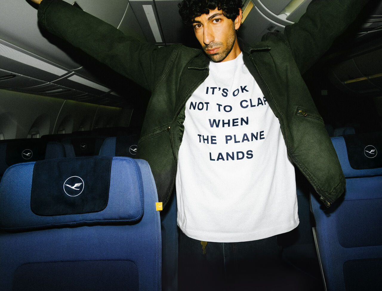A man stands in the aisle of a Lufthansa plane, wearing a green jacket and a white t-shirt that reads "IT'S OK TO CLAP WHEN THE PLANE LANDS"