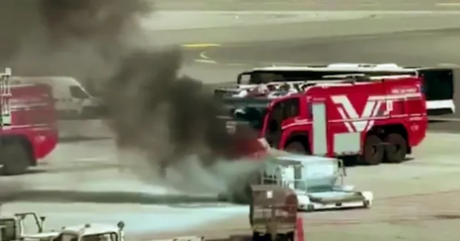 in the centre right of the image, a grey container can be seen with a large cloud of grey and black smoke billowing out from it towards the upper left corner. Behind and beside the container at a short distance are red emergency services vehicles arriving to help.