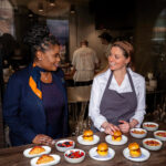JetBlue representative and chef stand with various plates of food in a a busy restaurant scene.