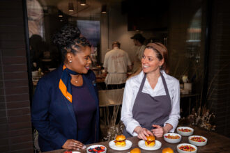 JetBlue representative and chef stand with various plates of food in a a busy restaurant scene.