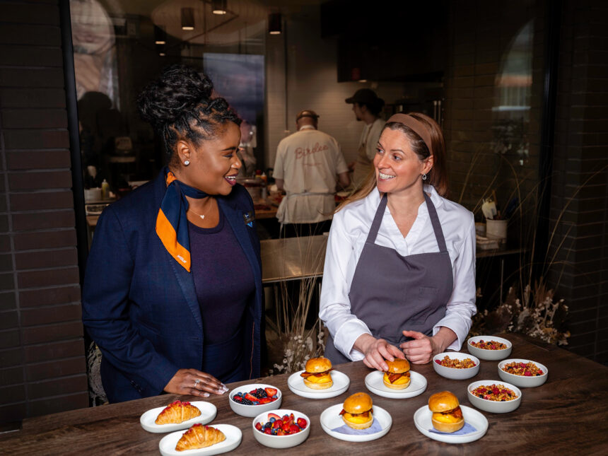 JetBlue representative and chef stand with various plates of food in a a busy restaurant scene.