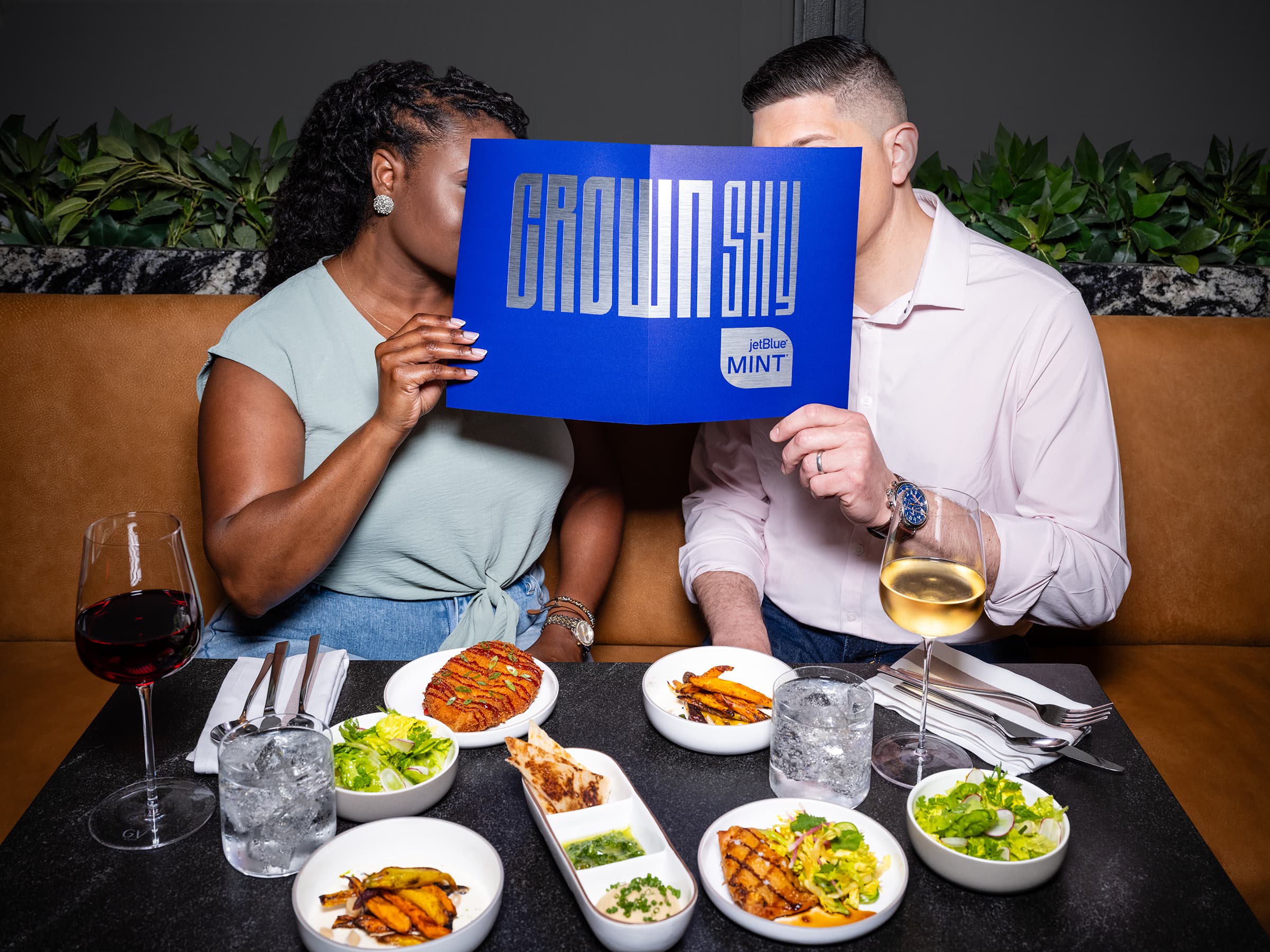 lady and gentleman sit at a restaurant table with lots of dishes holding a Crown Shy menu up to their faces, covering them.