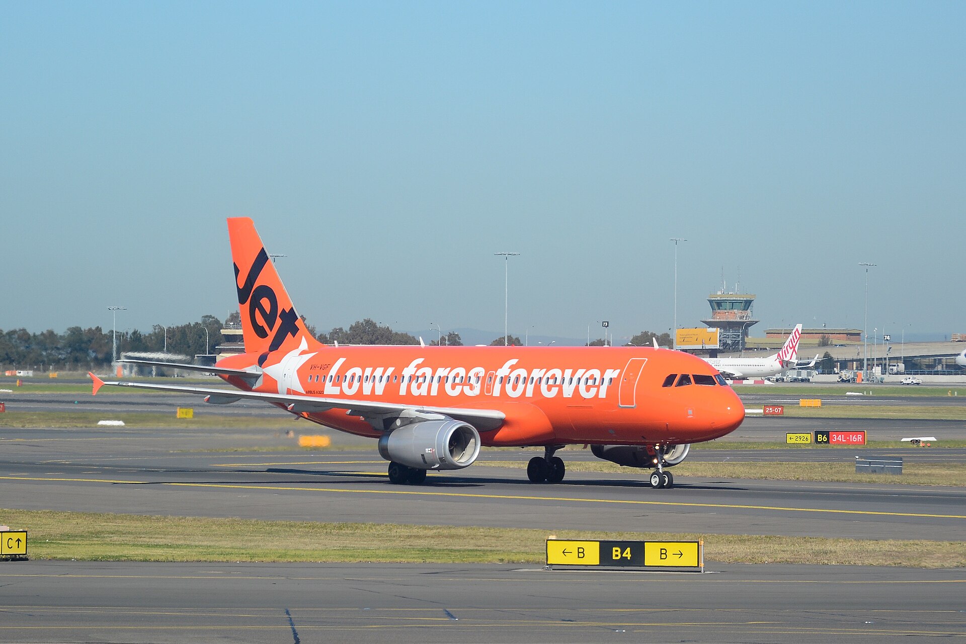 Jetstar A320 on Sydney Airport runway. The orange plane says "low fares forever" in white lettering.
