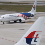 A Malaysia Airlines aircraft photographed in the background, positioned on an airport runway. At the forefront of the image is the tail of another aircraft, clearly displaying the Malaysia Airlines logo.