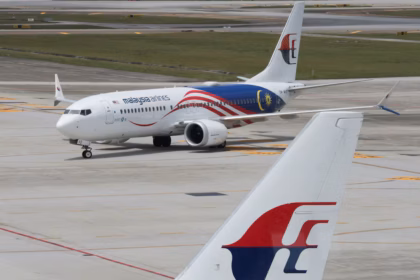 A Malaysia Airlines aircraft photographed in the background, positioned on an airport runway. At the forefront of the image is the tail of another aircraft, clearly displaying the Malaysia Airlines logo.