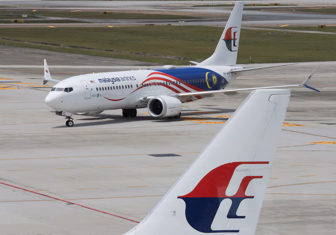 A Malaysia Airlines aircraft photographed in the background, positioned on an airport runway. At the forefront of the image is the tail of another aircraft, clearly displaying the Malaysia Airlines logo.