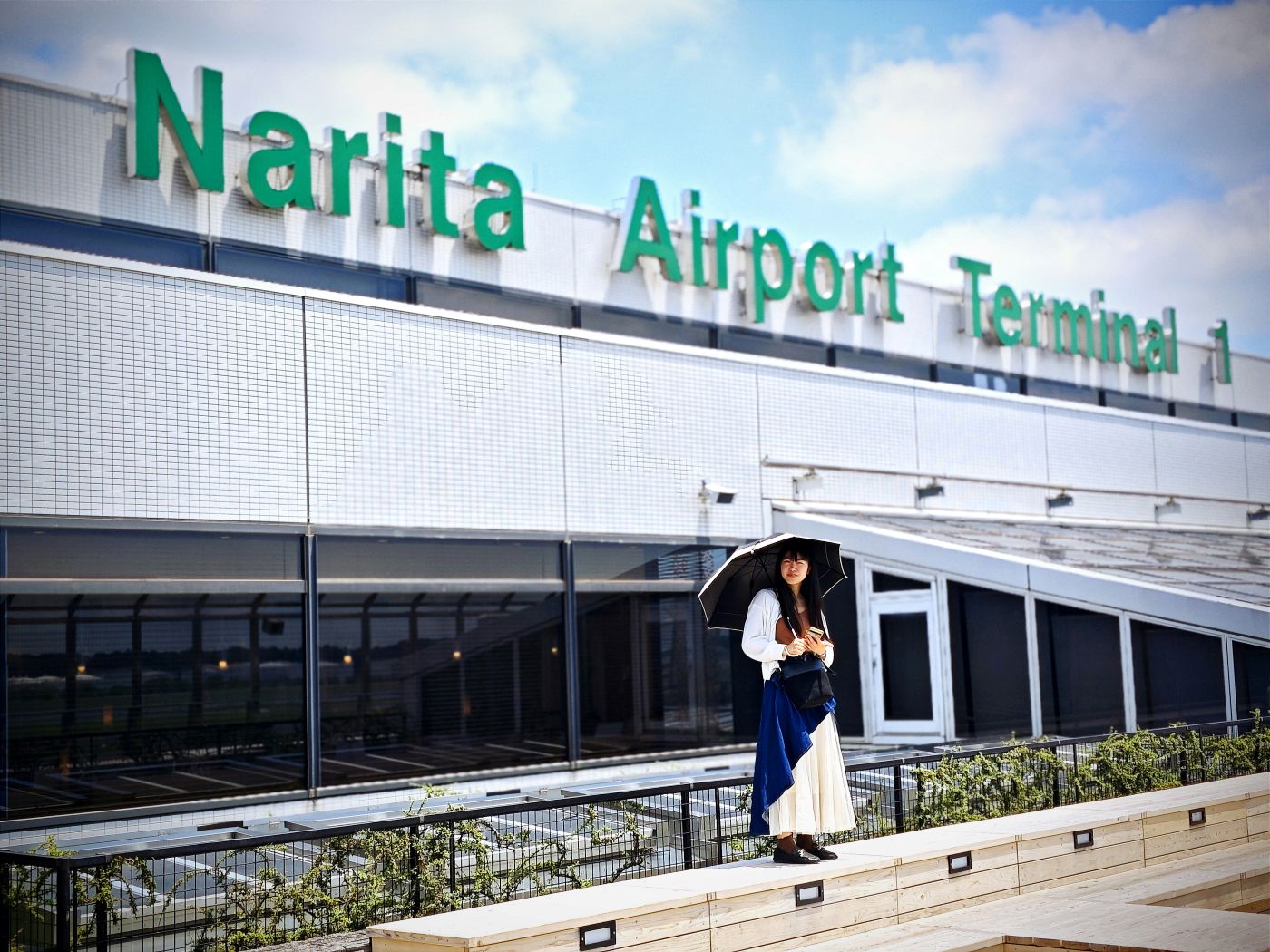 A front view of Narita International Airport terminal 1 with a cloudy blue sky backdrop and a woman holding an umbrella outside the door, dressed in white and blue.