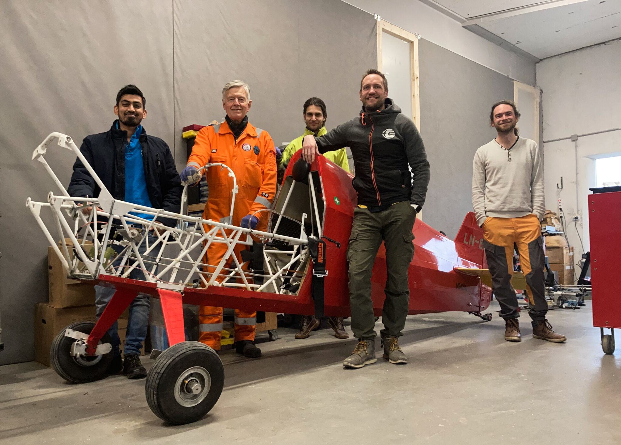 Five happy engineers/technicians are stood around the tail end of a electric sea plane that is not yet fully built. The unfinished aircraft is red with a white interior structure and two wheels.