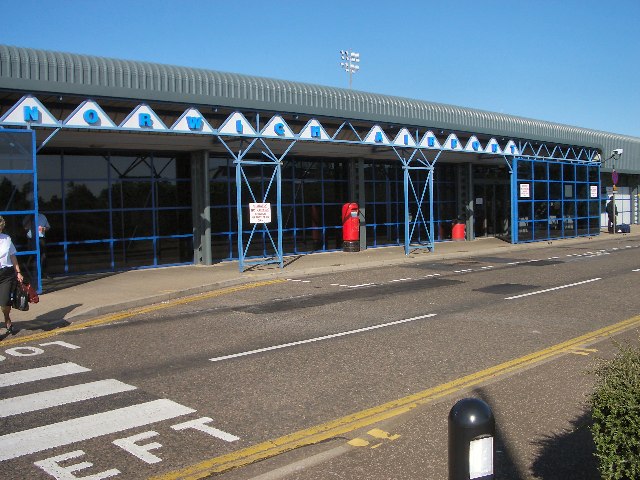 The entrance of Norwich Airport in 2005 taken at an angle. The walls are floor to ceiling windows divided up by blue frames. Pillars outside are blue, and stand beneath the sign which saying "Norwich Airport", each letter the sae blue in a white triangle. The top of the image shows cloudless blue sky. The foreground is a road with a zebra crossing visible in the bottom left corner.