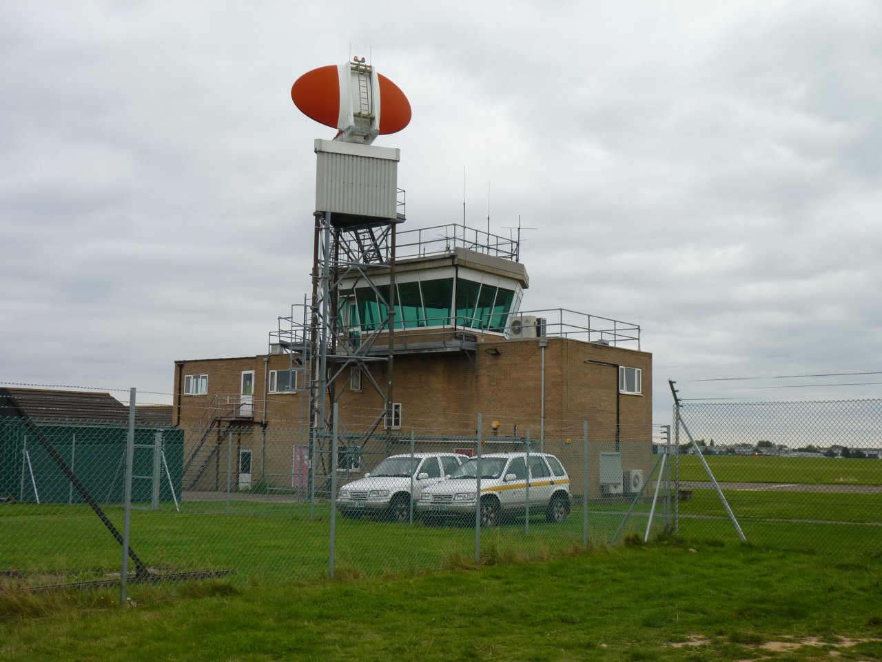 The control tower at Norwich Airport pictured from an angle. The base of the tower is a rectangular brick building in the centre of the image, with the glass watchtower part emerging from the centre of the top. In the foreground, grass leads up to the tower, which is wire fenced off, and two cars are parked in front. The background depicts grey cloudy sky.