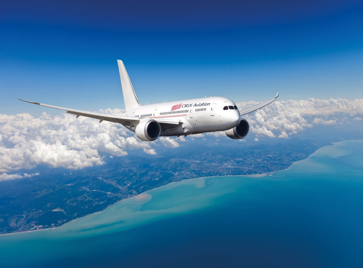 A ORIX Aviation plane in mid-flight with a deep blue sky and ocean background. The plane is bright white with the ORIX Aviation red logo. There are clouds off to the distance along with the edge of a country.