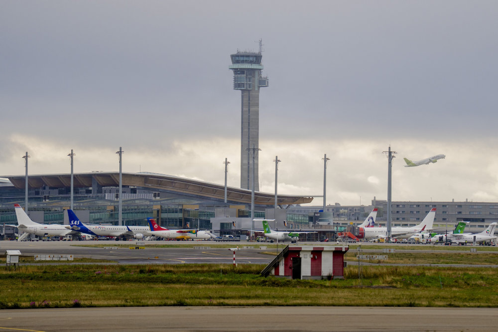 Oslo Gardermoen Airport runway pictured from afar, showing planes parked at gates and one taking off from the terminal.