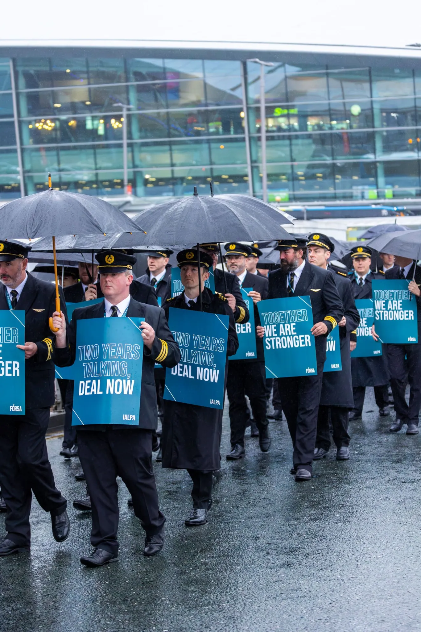 Aer Lingus pilots march in a long line at Dublin Airport. They are holding umbrellas in heavy rain and green signs, which read