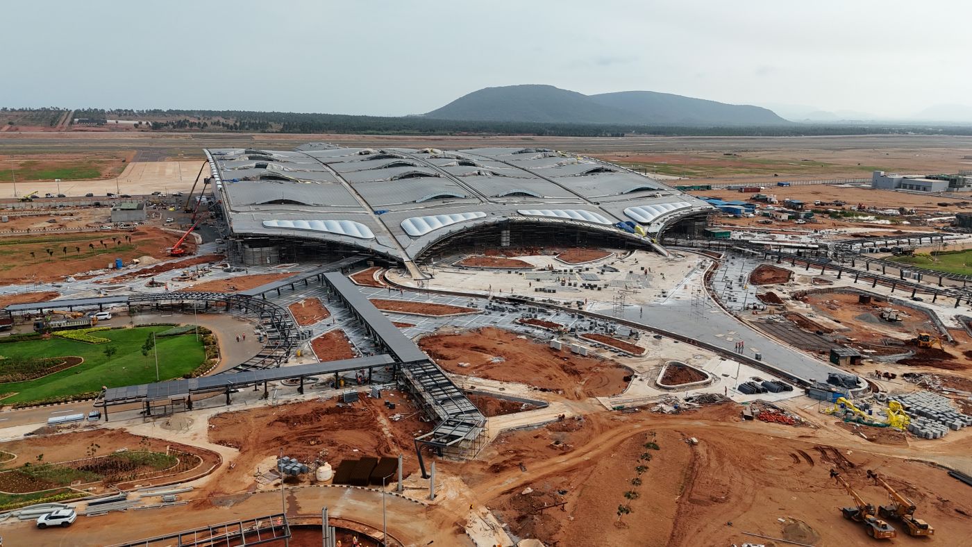 An aerial view of Bhopaguram Airport terminal under construction, showing patches of greenery surrounding the building's metal foundations and rooftop