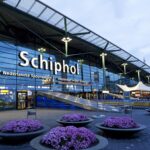 Exterior of the Amsterdam Airport Schiphol terminal entrance at night, illuminated signage above the doors and flower planters lining the walkway.