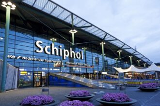 Exterior of the Amsterdam Airport Schiphol terminal entrance at night, illuminated signage above the doors and flower planters lining the walkway.