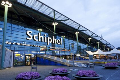 Exterior of the Amsterdam Airport Schiphol terminal entrance at night, illuminated signage above the doors and flower planters lining the walkway.
