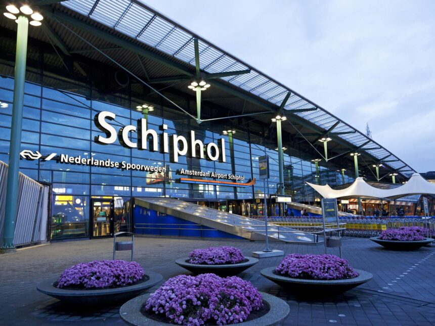 Exterior of the Amsterdam Airport Schiphol terminal entrance at night, illuminated signage above the doors and flower planters lining the walkway.