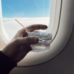 A left hand holding a small, plastic cup of water with ice cubes and a small, narrow straw in front of an aircraft window.