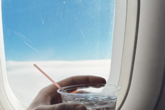 A left hand holding a small, plastic cup of water with ice cubes and a small, narrow straw in front of an aircraft window.