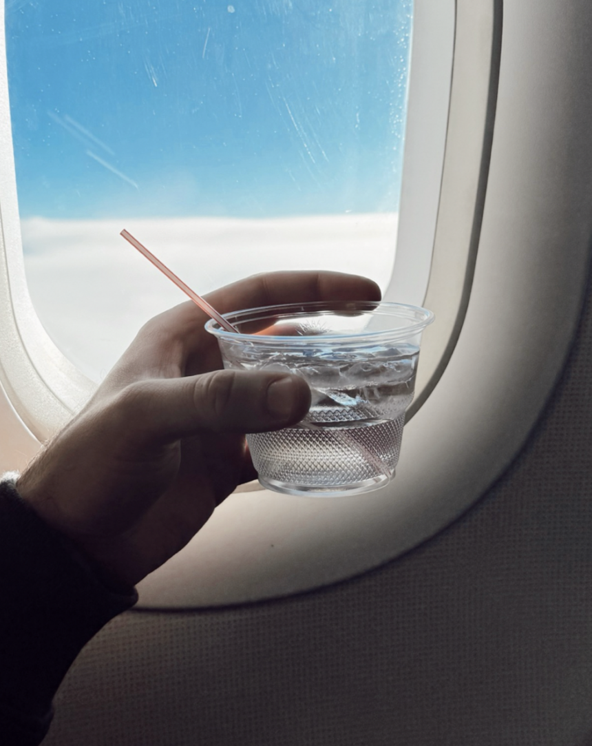 A left hand holding a small, plastic cup of water with ice cubes and a small, narrow straw in front of an aircraft window.