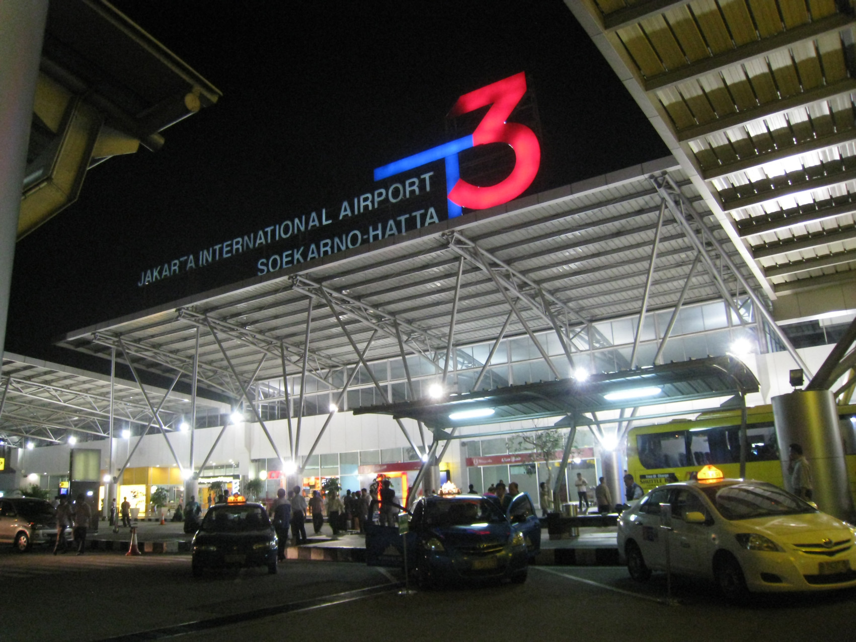 The taxi row outside Soekarno-Hatta International Airport Terminal 3 at night time, overlooking cars and a brightly lit terminal sign.