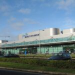 A front view of Belfast City Airport terminal with greenery outside and a clear blue sky background.