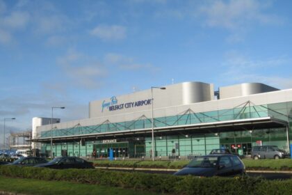 A front view of Belfast City Airport terminal with greenery outside and a clear blue sky background.