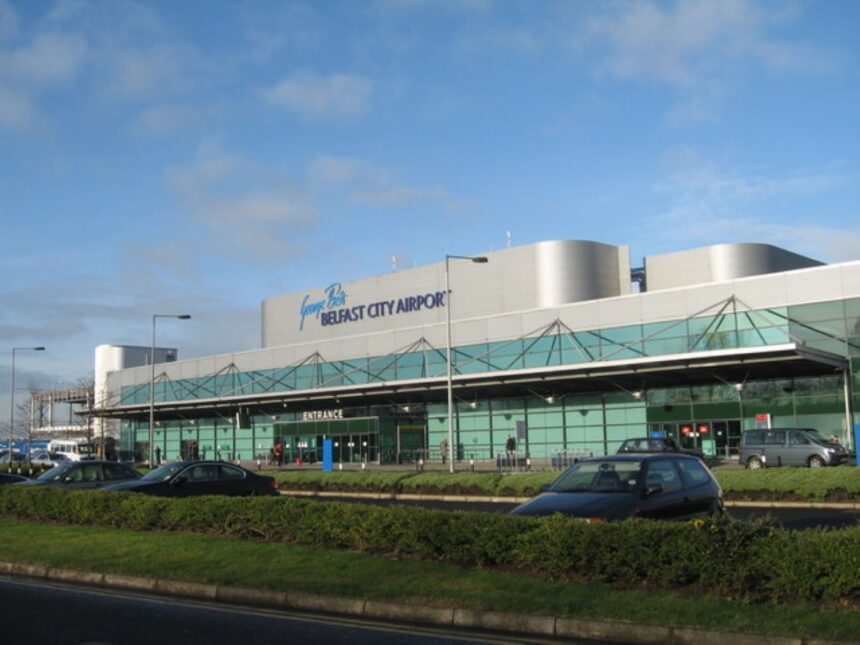 A front view of Belfast City Airport terminal with greenery outside and a clear blue sky background.