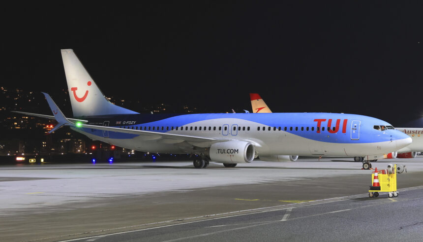 A TUI aircraft parked on the tarmac of an airport. A dark night sky is the background and the lights of various buildings and vehicles can be seen just behind the plane.