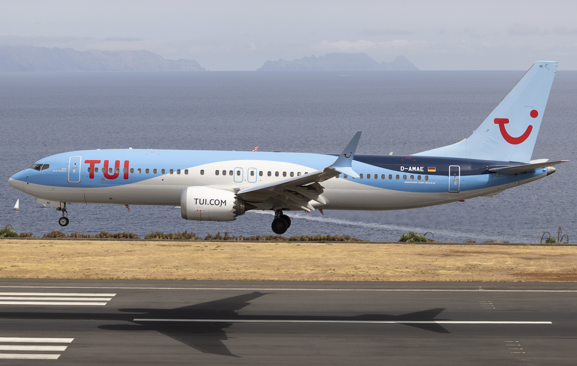 A TUI Boeing 737-MAX elevated slightly above the tarmac, with its shadow appearing on the runway beneath it. A body of water can be seen just behind the runway in the background.