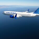 A United Airlines Boeing 737 Max-8 pictured flying over a body of open water against blue skies.