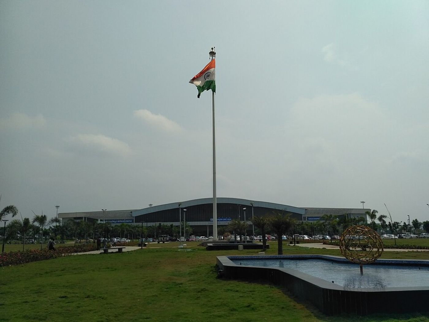A front view of Visakhapatnam Airport termminal with greenery and a lake and an India flag on a high mast in the front