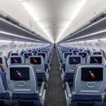 Interior view of a modern commercial aircraft cabin with multiple rows of blue economy-class seats arranged on either side of a central aisle. Each seatback is equipped with an in-flight entertainment screen displaying the American Airlines logo. Overhead baggage bins are closed, and bright cabin lighting creates a clean, symmetrical appearance extending toward the rear of the aircraft