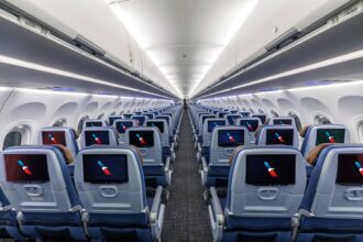 Interior view of a modern commercial aircraft cabin with multiple rows of blue economy-class seats arranged on either side of a central aisle. Each seatback is equipped with an in-flight entertainment screen displaying the American Airlines logo. Overhead baggage bins are closed, and bright cabin lighting creates a clean, symmetrical appearance extending toward the rear of the aircraft