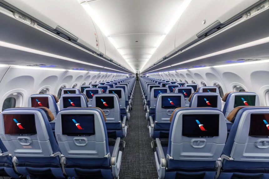 Interior view of a modern commercial aircraft cabin with multiple rows of blue economy-class seats arranged on either side of a central aisle. Each seatback is equipped with an in-flight entertainment screen displaying the American Airlines logo. Overhead baggage bins are closed, and bright cabin lighting creates a clean, symmetrical appearance extending toward the rear of the aircraft