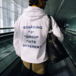 A person stands with their back to the camera, standing on an airport escalator. On their hoodie it reads "BOARDING BY GROUP HITS DIFFERENT"