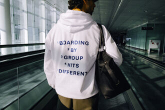 A person stands with their back to the camera, standing on an airport escalator. On their hoodie it reads "BOARDING BY GROUP HITS DIFFERENT"