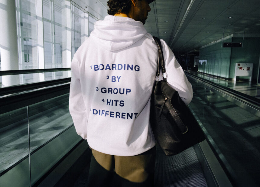 A person stands with their back to the camera, standing on an airport escalator. On their hoodie it reads "BOARDING BY GROUP HITS DIFFERENT"