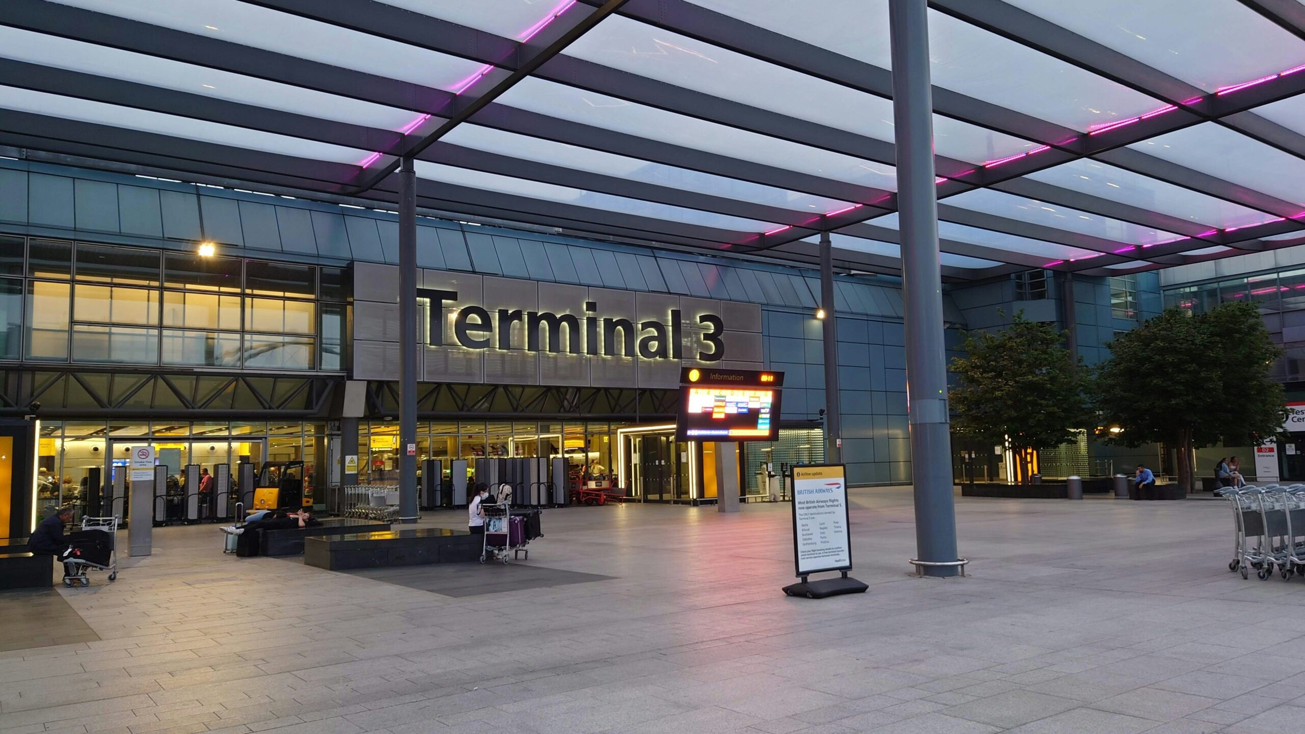 Airport Terminal 3 entrance with illuminated signage, glass canopy, information display, and luggage trolleys outside
