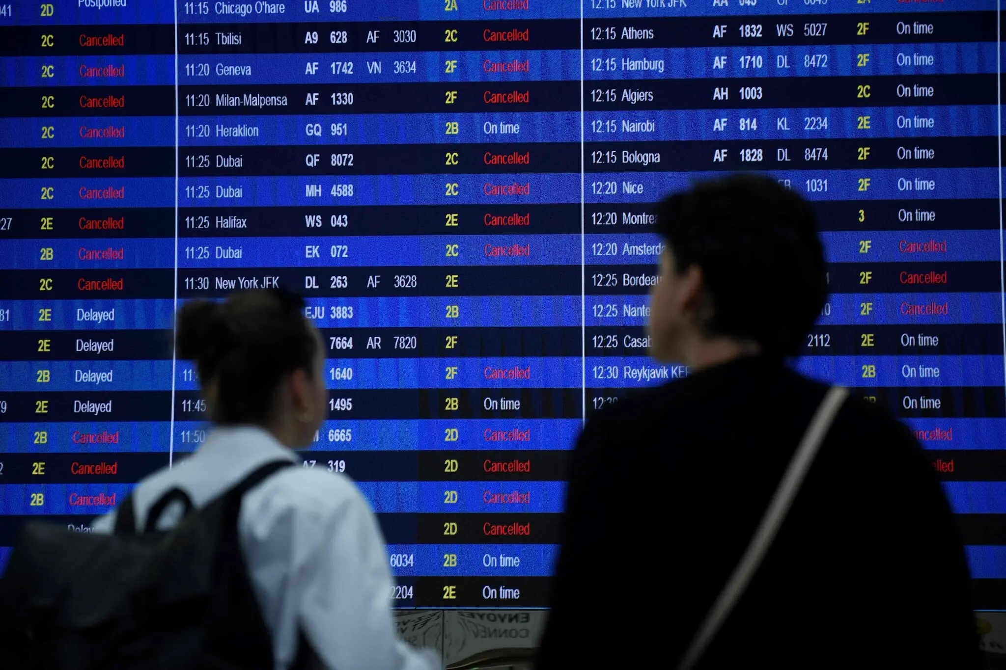 Two travellers stand on front of a departures board in Paris Charles de Gaulle Aiport (CDG) , staring at a long lost of cancelled and delayed flights.