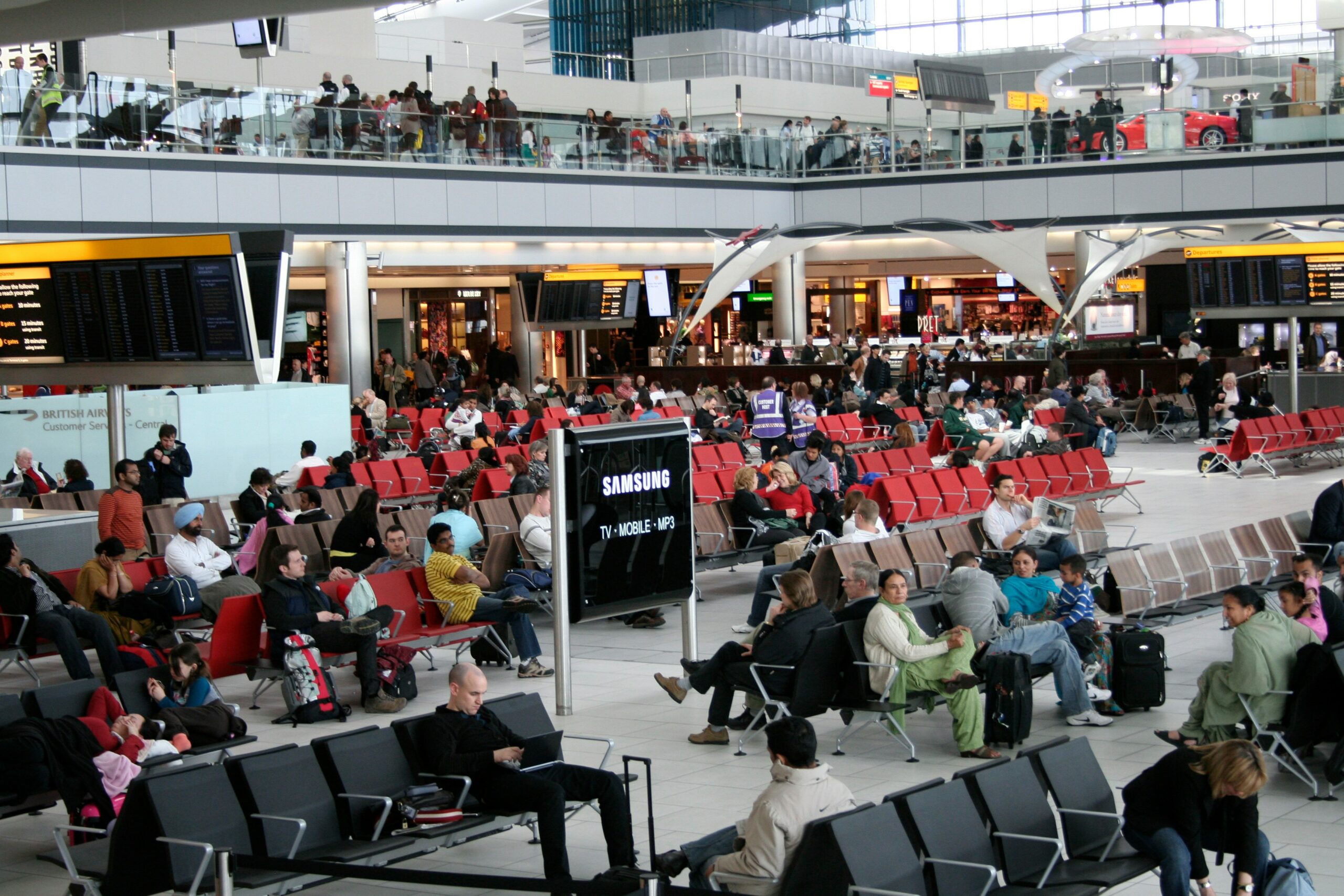 Busy airport departure lounge with rows of red seats, passengers waiting, and flight information screens overhead