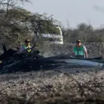 A picture taken from the crash site of the tourist plane flying over the Nazca Lines. A burnt part of the aircraft can be seen with two men stood behind it. A vehicle can be seen in the background.